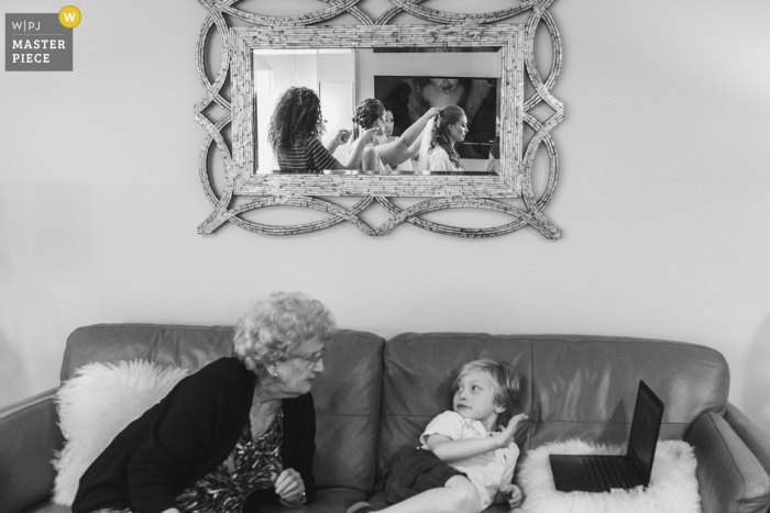 The bride is seen in the mirror on the wall getting ready for her wedding, while a child and older woman sit on a sofa with a laptop