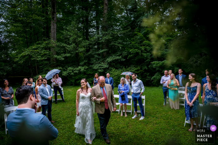 La sposa è scortata lungo il corridoio da suo padre, la prospettiva è dall'officiante, lo sposo si tiene la mano sul viso in soggezione al loro matrimonio nel cortile di Falls Church, Virginia