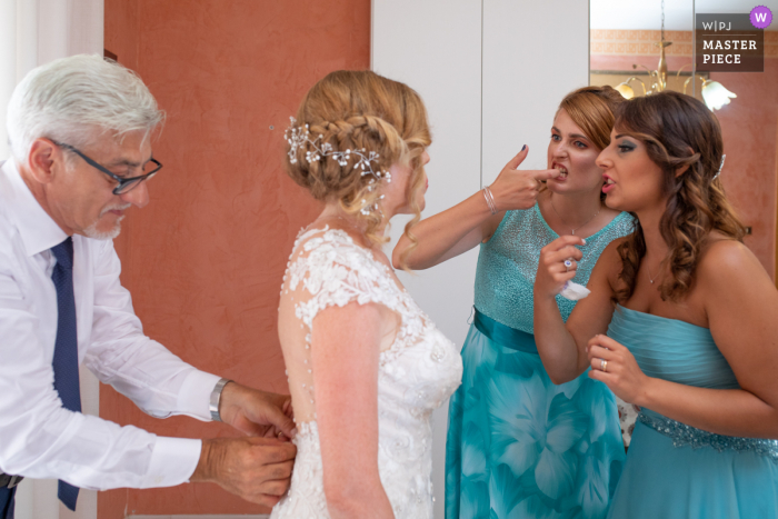 Siracusa bride receives help with her dress from her father and help from bridesmaids with something on her teeth