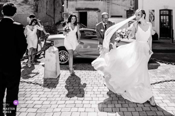 Fotografia francesa de casamento na Igreja de Poussan, no sul da França, mostrando a noiva caminhando em direção à entrada da igreja com um vento forte