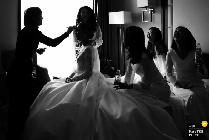 Fotografia di matrimonio in Cina nell'hotel di Pechino Prima della cerimonia, la sposa si sta rammendando il trucco e le damigelle ridono al gioco del matrimonio