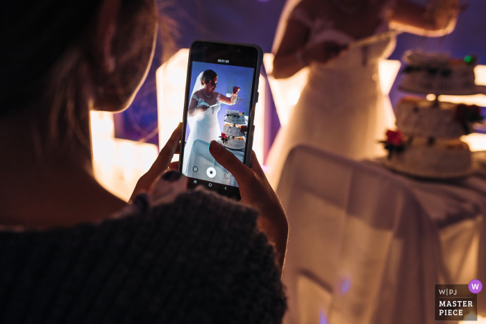 Un photographe de mariage de Lodzkie a créé cette image au Monastère des Pères Bernardins à Piotrkow Trybunalski, Pologne - Une photo par-dessus l'épaule du téléphone de l'invité du mariage filmant la mariée en train de couper un gâteau