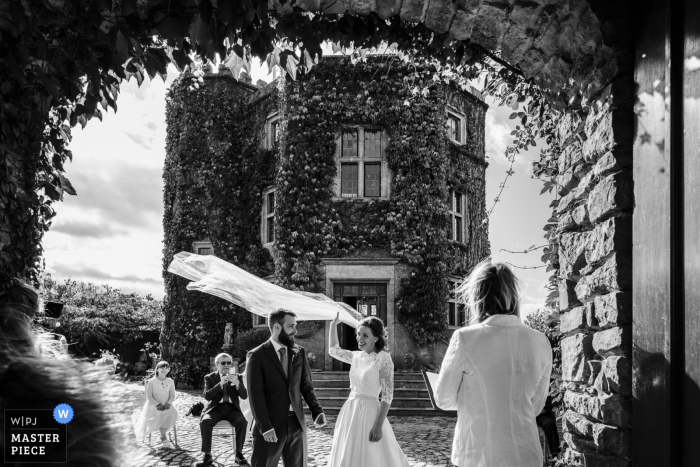 Fotografia di reportage di matrimonio nel Regno Unito da Walton Castle, Somerset, Inghilterra che mostra il velo della sposa sollevato dal vento durante la cerimonia nuziale - Immagine incorniciata da un arco nelle mura del castello