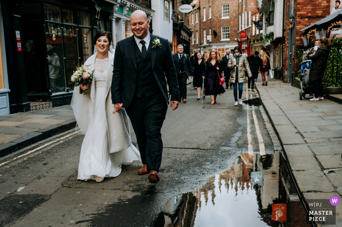 Imagem do casamento da noiva e do noivo caminhando alegremente pelas ruas de York, com a Catedral refletida em uma poça