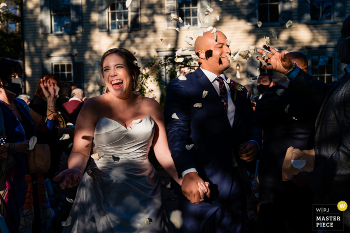 Fotografía de boda de PA al aire libre de Filadelfia del novio con flores arrojadas en su cara mientras él y su ahora esposa dejan la ceremonia en un día muy soleado