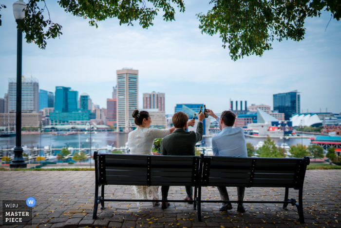 Fotografía de boda que muestra a la pareja y al hermano de la novia vitoreando con su mini-champán en el parque con vistas al puerto interior de Baltimore