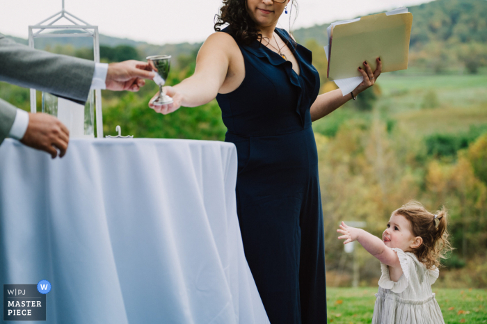 De huwelijksfotograaf van het District Of Columbia heeft deze foto gemaakt in The Inn at Mount Vernon Farm, Sperryville, VA van een klein bloemenmeisje dat om een ​​glas wijn vroeg tijdens de Joodse ceremonie