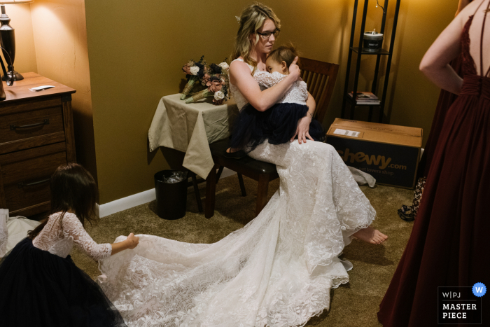 Foto van een bruid uit Schenectady, NY die haar bruidsjurk draagt, houdt haar slapende baby vast terwijl een klein bloemenmeisje haar sleep rechtzet.