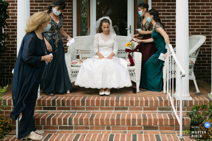 Immagine di una sposa seduta su una sedia di vimini in tranquilla contemplazione mentre aspetta di incontrare il suo sposo, mentre le sue donne membri della famiglia si stabiliscono intorno a lei a Long Island, NY