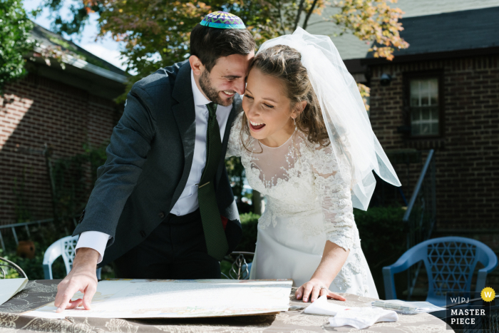 Huwelijksfoto in Long Island, NY van een A-bruid en bruidegom die vreugdevol dicht bij elkaar zitten terwijl ze voor het eerst hun huwelijksketubah bekijken.