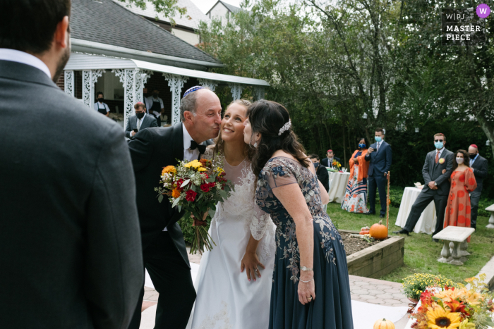 Foto que muestra a una novia besada en la mejilla por su madre y su padre al mismo tiempo que se la pasan al novio al comienzo de una ceremonia de boda al aire libre en Long Island, NY.