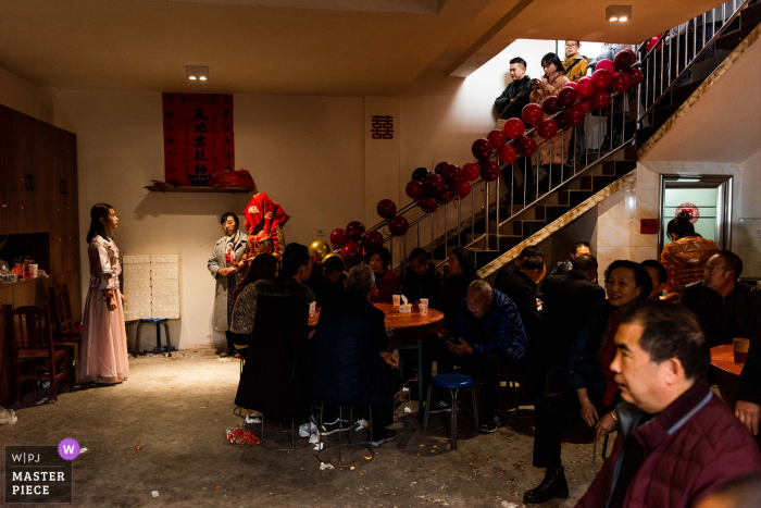 Fotografia de casamento na China de Sichuan na casa da noiva - sob a luz à esquerda está a mãe da noiva segurando a mão da noiva. A cabeça da noiva é coberta com um véu vermelho