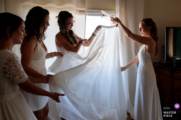 FR Fotografía de la boda de la casa de los novios mientras la novia despliega su velo con la ayuda de sus amigos.