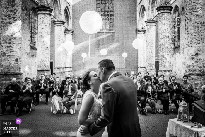 Foto de casamento de Oude Kerk, Dongen - o beijo durante a cerimônia, os convidados do casamento ao fundo