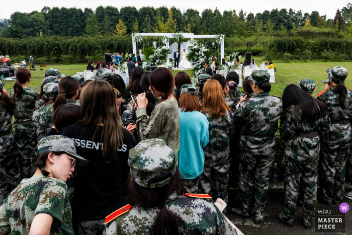 Trouwfoto uit China - De pasgetrouwden houden een huwelijksceremonie. Een groep militaire trainingsstudenten in de buurt kijkt naar de huwelijksceremonie.