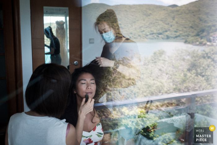 Fotografia ślubna z Wong Keng Tei, Sai Kung, Hong Kong (dom Panny Młodej) — panna młoda ma zrobiony makijaż, naturalne otoczenie jej domu i jej suknia ślubna odbijają się w szklanych drzwiach tarasu.