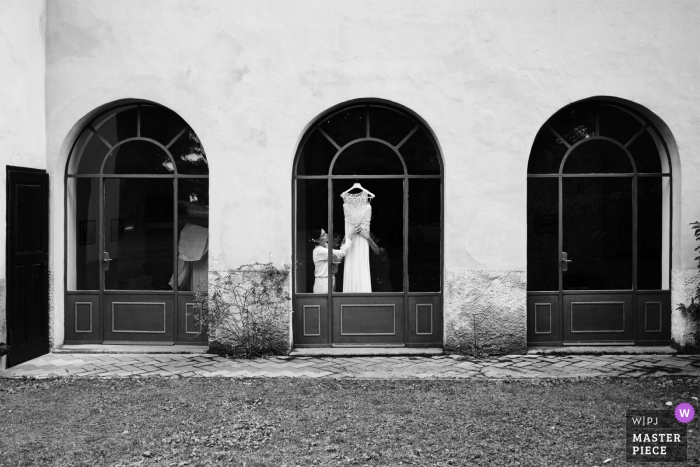 Huwelijksfotografie in Italië in zwart-wit van Villa Caproni, Vizzola Ticino van de voorbereiding met bruidsjurk die in een raam hangt