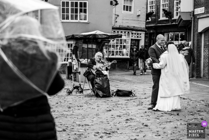 Het huwelijksbeeld van York City Center van de bruid en bruidegom die dansen op straat, in de regen, terwijl een straatmuzikant verder speelt