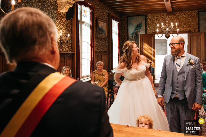 Flandern Hochzeitsfotografie in einem Rathaus des Sohnes des Brautpaares beobachtet den Minister während der Zeremonie genau unter dem Tisch