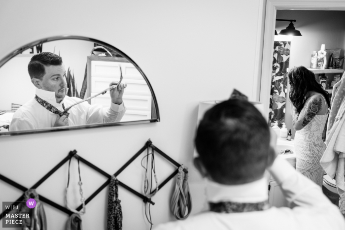 Fotografía de boda del novio trabajando en atarse la corbata mientras su novia se pone los pendientes en el baño. Se estaban preparando juntos para dirigirse a las montañas para su fuga con solo ellos dos y un oficiante.