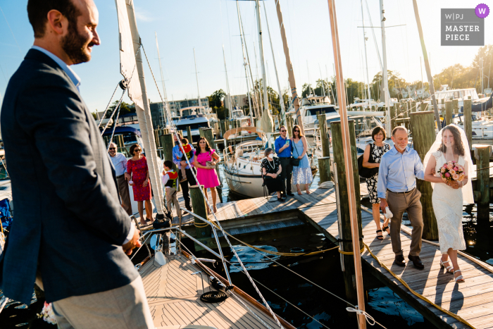 Hochzeitsfoto aus Pennsylvania des Bräutigams, der auf dem Segelboot seiner Familie auf seine Braut wartet, die mit ihren Eltern das Dock herunterkommt, während sie auf dem Segelboot heiraten wollen