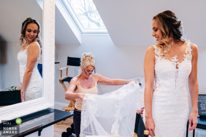 Hochzeitsfotografie von West Sussex aus der Brautvorbereitungssuite in der Brookfield Barn in Horsham Bride mit dem reflektierten Spiegel, während die Mutter der Braut ihr Kleid anpasst