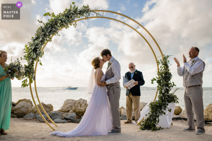 Ein Hochzeitsfotograf in Key West, Florida, im Fort Zachary Taylor Park hat dieses Bild des vorbeifahrenden Paares mit seiner Liebe zu Booten erstellt, als sie sich bei der Zeremonie im Freien zum ersten Mal als Ehemann und Ehefrau küssten