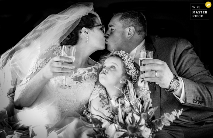 Beste Hochzeitsfotografie aus Wicklow, Irland, die ein Bild von einem echten, aufrichtigen Moment im Auto mit dem Paar und einem Kind während eines privaten Kusses mit einem Toast zeigt