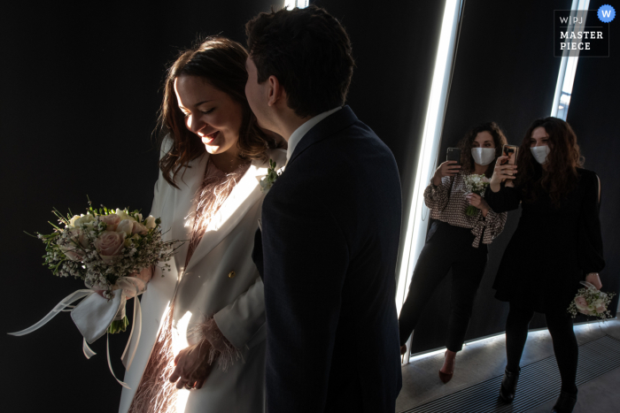 Un photographe de mariage à Quistello, Mantoue a créé cette image au Centro Hub Oltrepo Mantovano des mariés vont à la sortie, pendant que le marié murmure quelque chose à l'oreille de la mariée, Pendant ce temps, les témoins recherchent une photo