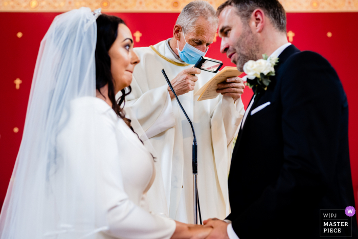 A top Ireland wedding reportage photographer at the CloughJordan House captured this picture showing Priest needs a little help reading the vows during the ceremony