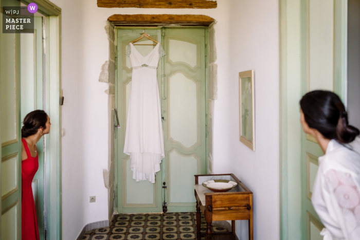 Un photographe de mariage du Domaine Fon de Rey dans l'Hérault a créé cette image de la mariée et de la demoiselle d'honneur inspectant la robe de mariée suspendue au-dessus d'une porte