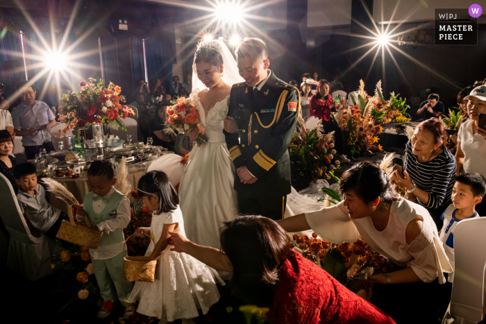 A melhor fotografia de casamento de Sichuan mostrando uma foto mostrando O noivo e a noiva caminhando em direção ao palco. Na frente deles estão duas crianças carregando cestos de flores. As pessoas no canto inferior direito dizem às crianças para andarem mais rápido