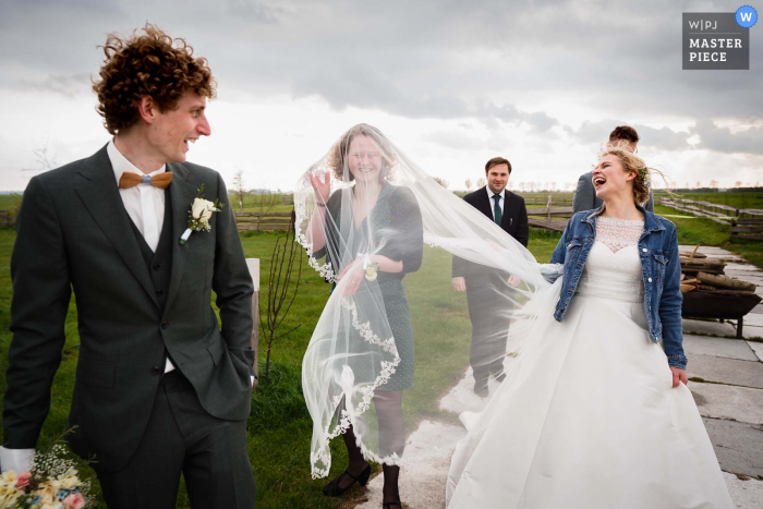 Een top huwelijksfotograaf in Nederland heeft deze foto gemaakt en laat zien dat het die dag winderig was en de vriend van de bruid had een grappig moment met de sluier die opsteeg door de wind