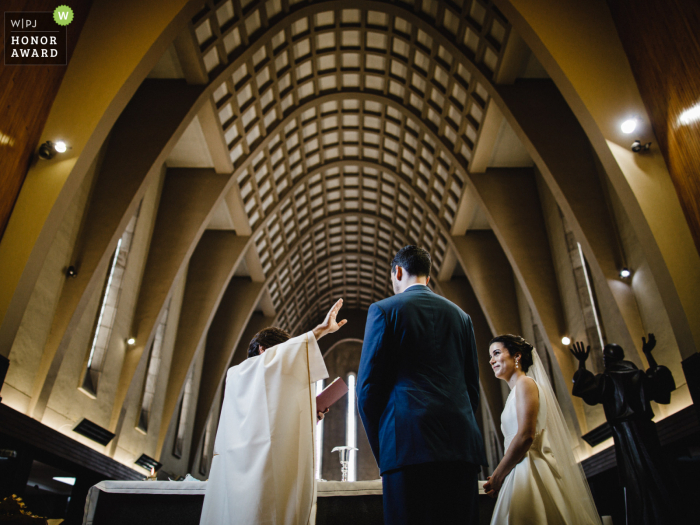 Preisgekröntes Bildbeispiel des Hochzeitsfotografenwettbewerbs in Porto, Portugal, das den Priester mit erhobenen Händen zeigt, der Braut und Bräutigam in der Kirche segnet