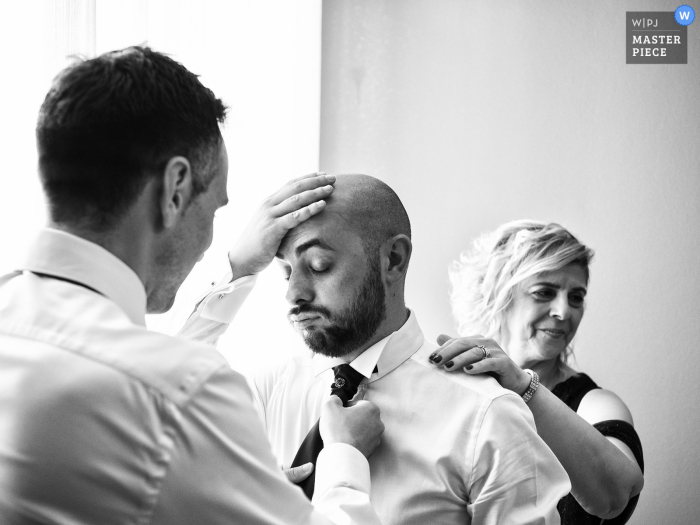 Best wedding photography from Milan showing The mother and the best man helping him fix the tie but at that moment the tension is felt and the groom cant take it anymore 