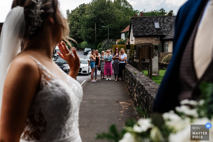 Uma igreja em East Yorkshire, Reino Unido, fotografia de reportagem de casamento mostrando a noiva acenando para uma multidão que a esperava quando ela chega para a cerimônia