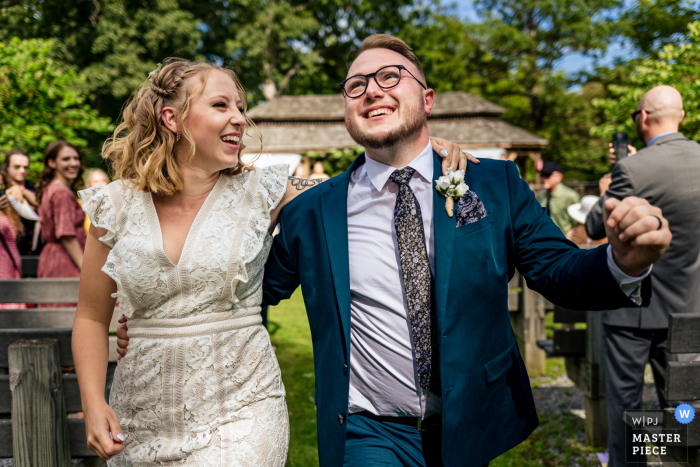 Imagem premiada da cerimônia de casamento ao ar livre no Parque Estadual Cacapon, West Virginia, mostrando o casal saindo alegremente sob as árvores e o céu azul. Os melhores concursos de fotos de casamento do mundo apresentados pela WPJA