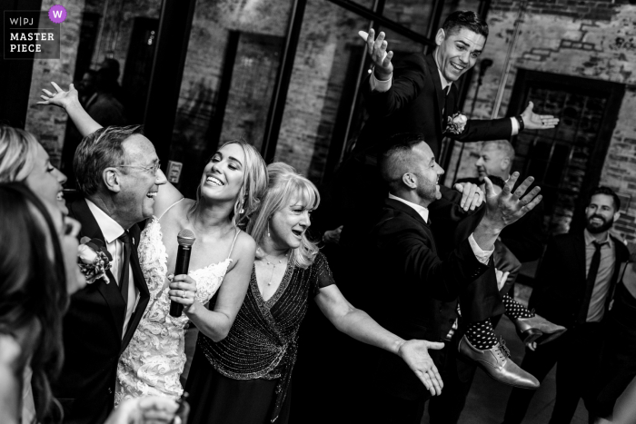 Mt. Washington Mill Dye House, Baltimore indoor wedding reception party award-winning picture showing Bride and parents singing at the venue while groom is lifted up in the air. The world's most skilled wedding photographers are members of the WPJA