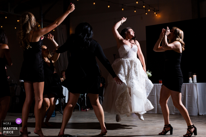 Clay Venues, Colorado Springs, bekroonde foto van een indoor bruiloftsreceptie waarop de bruid en haar besties springen van vreugde tijdens het vieren bij de receptie. 'S Werelds meest bekwame trouwfotografen zijn lid van de WPJA