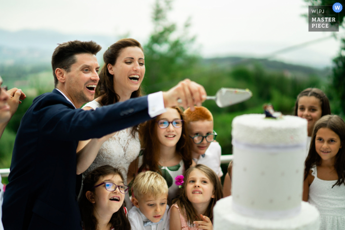 Tenuta Colle Paradiso, Erbusco Outdoor-Hochzeitsempfangsparty preisgekröntes Foto, das das Anschneiden der Hochzeitstorte im Kreise der kleinen Gäste festhält. Die besten Hochzeitsfotografen der Welt messen sich bei der WPJA