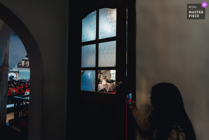 Imagen premiada de la ceremonia de matrimonio en interiores de Fujian que muestra a la dama de honor usando su teléfono móvil para grabar al recién llegado en la ceremonia desde la esquina.