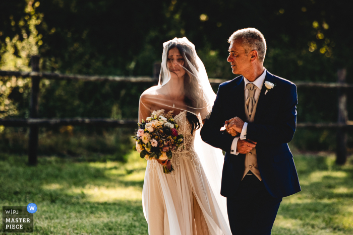 Imagem premiada da cerimônia de casamento ao ar livre Tenuta Pantano Borghese- Roma, mostrando a noiva entrando na cerimônia com o pai. Os melhores concursos de fotos de casamento do mundo apresentados pela WPJA