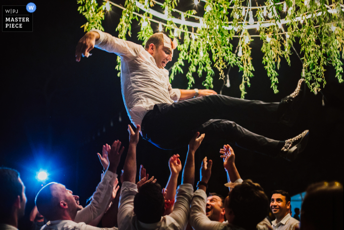 Bodrum, Turkey outdoor marriage reception party award-winning photo that has recorded a Groom in air under trees and lights. The world's top wedding photographers compete at the WPJA