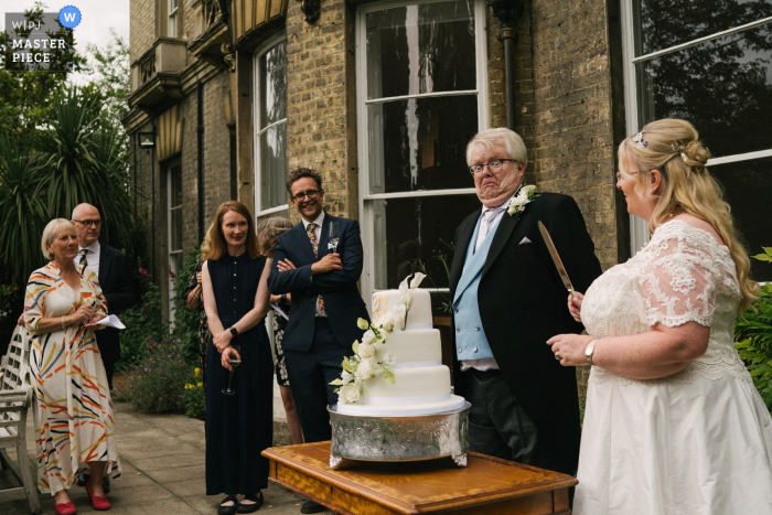 Bekroonde foto van een huwelijksreceptie in Cambridge, VK waarop een reactie van de bruidegom is vastgelegd op de bruid die het taartmes vasthoudt. 'S Werelds beste trouwfotografen strijden tegen de WPJA