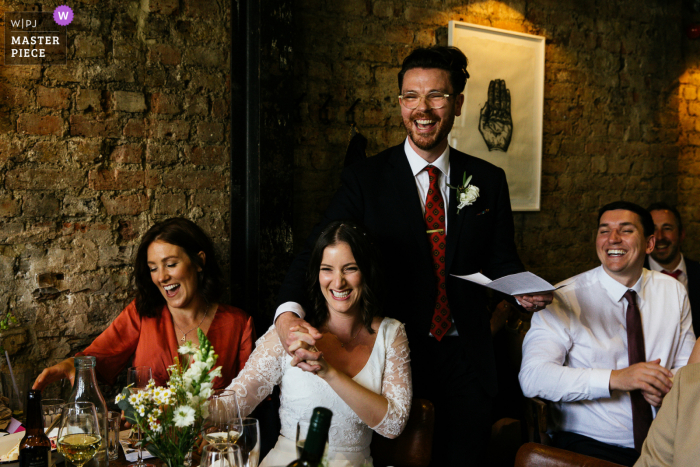 De bekroonde foto van de Elgin, London indoor bruiloftsreceptie waarop de bruidegom zijn toespraak houdt terwijl hij de hand van zijn glimlachende vrouw vasthoudt. 'S Werelds meest bekwame trouwfotografen zijn lid van de WPJA