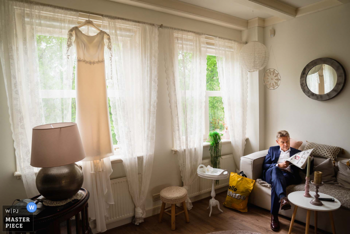 Castle Cammingha in Bunnik marriage preparation time award-winning picture capturing The father of the bride reading his newspaper while the wedding dress is hanging in the window.