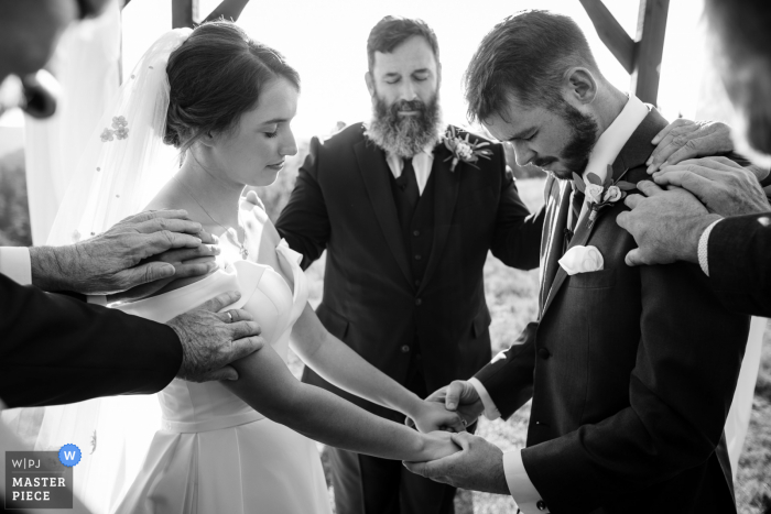 Imagem premiada da cerimônia de casamento ao ar livre de Grass Range, Montana mostrando os homens colocando as mãos no casal durante a oração. Os melhores concursos de fotos de casamento do mundo apresentados pela WPJA