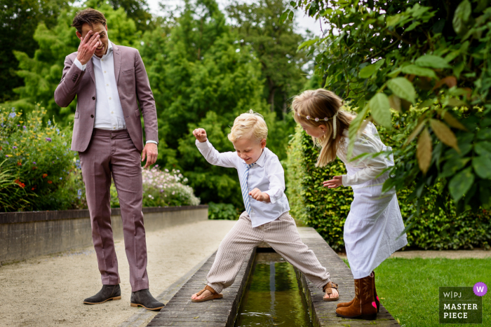 Noord-Brabant-Hochzeitstag preisgekröntes Bild eines jungen Brautmannes, der über Wasser springt, als die Brautjungfer ihn überprüft, der Vater runzelt die Stirn - von den weltweit besten Hochzeitsfotografie-Wettbewerben, die von der WPJA veranstaltet werden