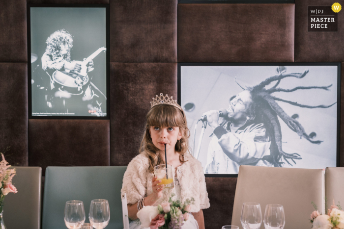 Foto pluripremiata del ricevimento di matrimonio di Frederick's, Camden, Londra, che ha registrato la fioraia del matrimonio sorseggiando un drink con un'espressione innocente, sullo sfondo di immagini iconiche del mondo della musica popolare