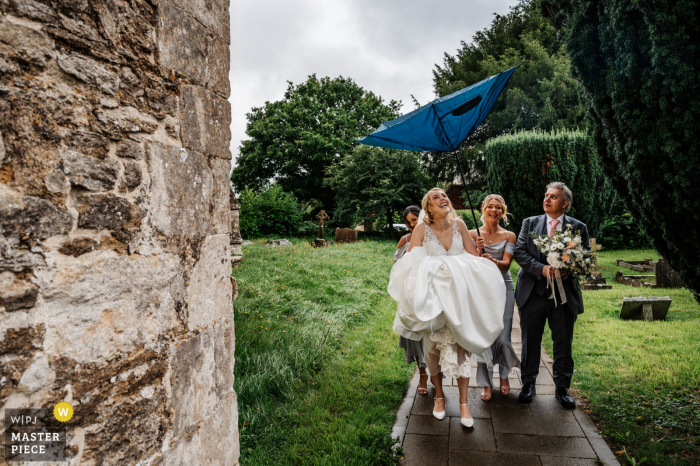 Somerset, Inghilterra, immagine premiata per il giorno delle nozze della festa nuziale che arriva in chiesa sotto la pioggia mentre il vento soffia l'ombrello al rovescio - dai migliori concorsi di fotografia di matrimonio al mondo ospitati dal WPJA